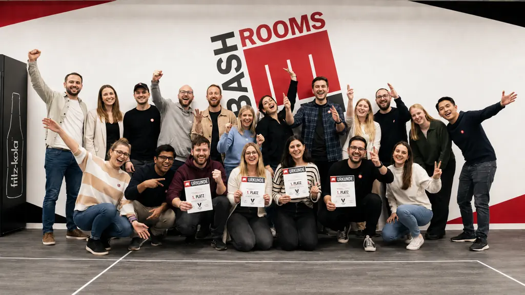 Große Gruppe von rund zwanzig Gästen vor der BASH-ROOMS-Wand, die vorderen knien mit Urkunden in der Hand