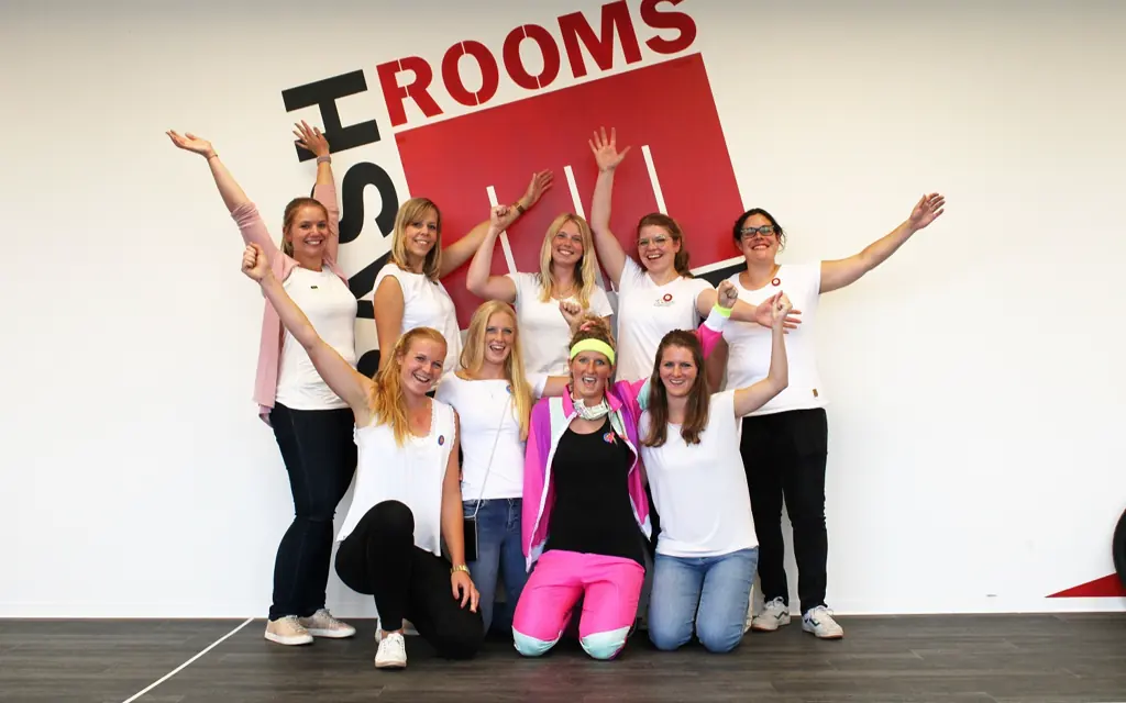 Zehn Frauen in weißen Shirts jubeln, die Braut in der Mitte trägt ein pinkes Stirnband