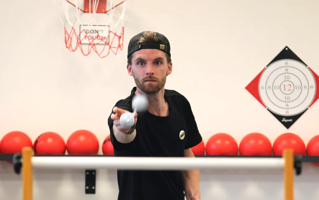 Ein Spieler in schwarzem Shirt und Kappe zielt mit einem Tischtennisball auf die Kamera, dahinter eine Reihe roter Bälle und eine Zielscheibe
